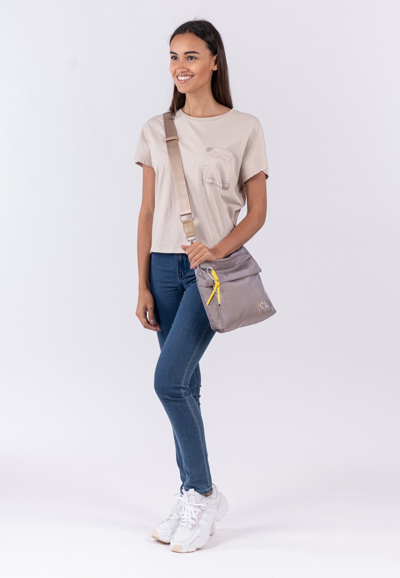 Borsa a tracolla grigia con zip gialla, texture in tessuto e design a ribalta. Indossata con una maglietta beige casual e jeans blu.