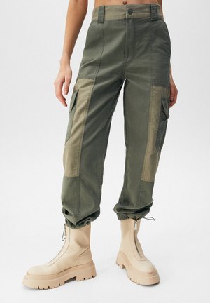 Pantalon cargo bicolore vert avec multiples poches, poignets élastiques, associé à des bottes beige épaisses avec fermeture éclair à l’avant, sur fond blanc.