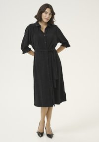 Robe noire à boutons avec manches courtes bouffantes, tissu texturé, ceinture à la taille et longueur au genou. Associée à des talons aiguilles noirs.