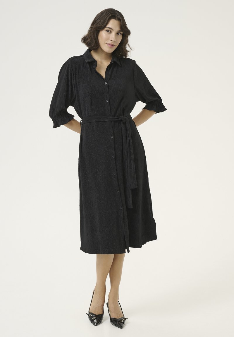 Robe noire à boutons avec manches courtes bouffantes, tissu texturé, ceinture à la taille et longueur au genou. Associée à des talons aiguilles noirs.