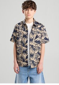 Camisa de manga corta en beige con estampado de hojas tropicales en azul marino, con cuello, cierre de botones y un bolsillo en el pecho. Se lleva sobre una camiseta blanca.