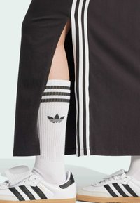 Baltas un melns Adidas Samba apavi, melni un baltu svītru augstās zeķes līdz ceļiem, un melna svārka ar baltām sānu svītrām.