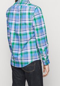 Osoba oblečená v dlouhém kostkovaném tričku v modré, zelené a bílé barvě, spárovaná s tmavými džínami, zobrazená zezadu na jednoduchém pozadí.