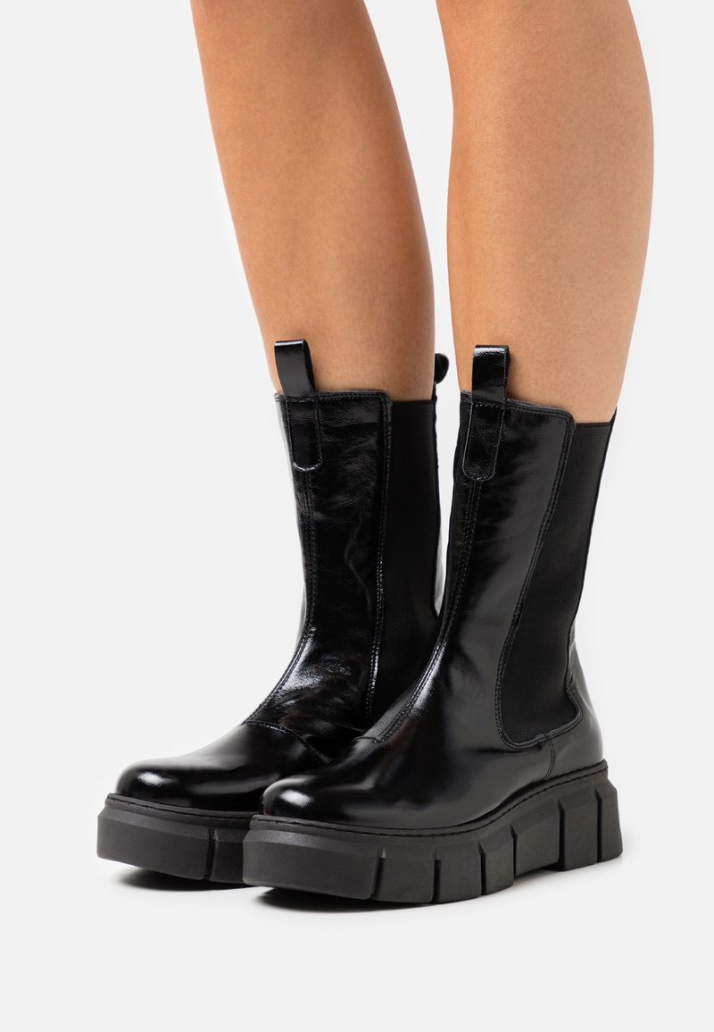 Bottes mi-mollet en cuir noir brillant avec semelles épaisses en caoutchouc portées sur des jambes nues sur un fond uni.