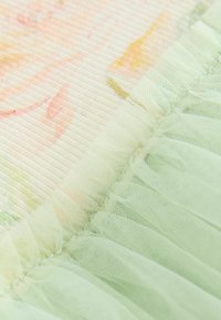 Gros plan sur un tissu vert menthe transparent à volants superposé à un textile à motif floral avec des tons pastel doux et une texture fine côtelée.
