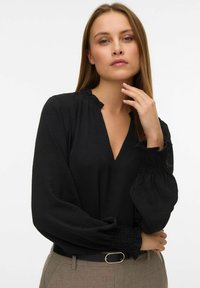 Melnas blūzes ar V-veida kakla izgriezumu, ruffled apkakli un garām spārnu piedurknēm ar smocked aprocēm. Viegls teksturēts audums. Bez raksta.