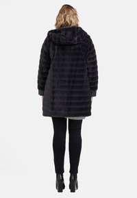 Czarny płaszcz z kapturkiem z imitacji futra o pikowanej fakturze, wykończony długimi rękawami i luźnym krojem, zestawiony z czarnymi spodniami o wąskim kroju i botkami na obcasie.