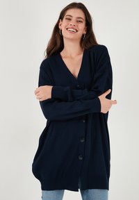 Tamsiai mėlynas kardiganas, pagamintas iš minkštos mezgimo medžiagos, su gilia V formos iškirpte, ilgomis rankovėmis ir šešiais dideliais mygtukais priekyje.