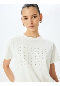 Valkoinen lyhythihainen t-paita, jonka etuosa on teksturoitu ja siinä on pieniä helmiä ja timantteja vaihtelevissa kooissa muodostamassa ruudukon.