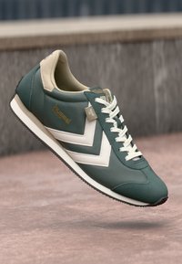 Sneaker sintetico verde con accenti beige, presenta un design a chevron bianco, punta tondeggiante, suola a contrasto e allacciatura frontale. Dettaglio del marchio in oro.