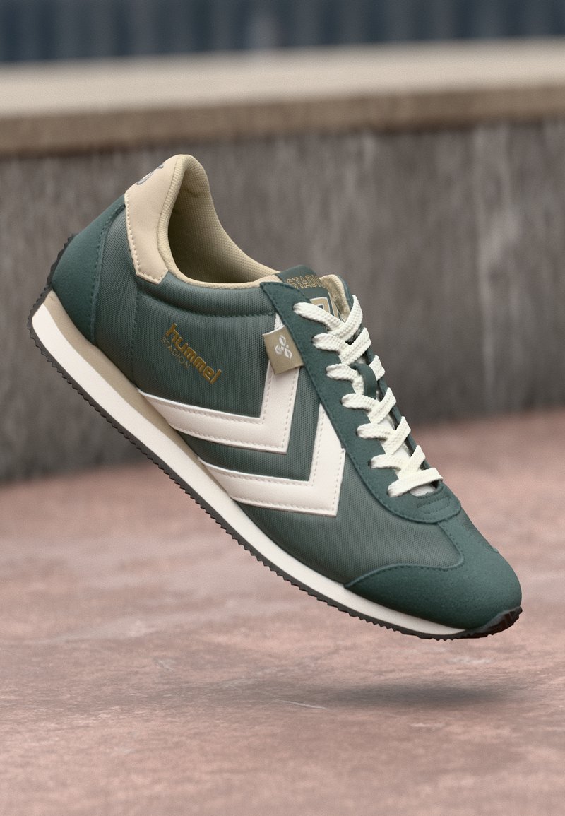 Sneaker sintetico verde con accenti beige, presenta un design a chevron bianco, punta tondeggiante, suola a contrasto e allacciatura frontale. Dettaglio del marchio in oro.
