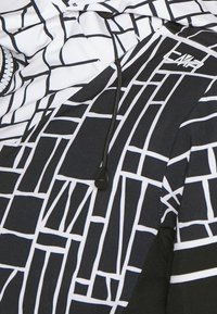 Veste à motifs noir et blanc avec un design en grille, dotée d'une capuche à cordon de serrage et d'une fermeture éclair à l'avant. Texture douce avec des sections de tissu contrastantes.