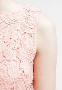 Detailní záběr na světle růžový bezrukávový oděv s květinovou krajkovou výšivkou na bledé paži na bílém pozadí.