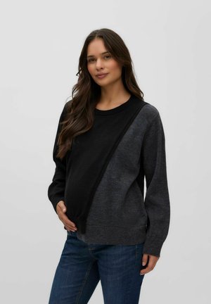 Femme enceinte aux longs cheveux bruns, portant un pull bicolore noir et gris et un jean bleu, tenant doucement son ventre.