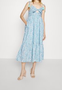 Blå blommig klänning med knyt fram, justerbara axelband och en lagad kjol. Har fickor och är parat med nude blockklacksandaler.