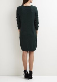 Kvinna i en mörkgrön långärmad knälång klänning och svarta högklackade ankelstövlar, stående mot en helt vit vägg.