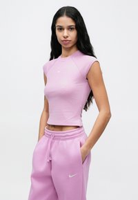 Γυναίκα με μακριά σκούρα μαλλιά, φορώντας ένα εφαρμοστό λιλά αμάνικο τοπ και αντίστοιχο παντελόνι φόρμας με λευκά λογότυπα Nike, στέκεται με τα χέρια στις τσέπες.