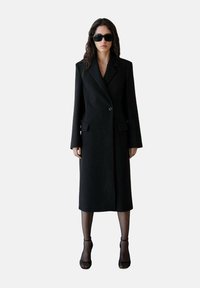 Lange zwarte jas van gestructureerde stof, met een enkele knoop, ingesneden revers en twee voorkruiszakken. Draag het over zwarte panty's en hakken.
