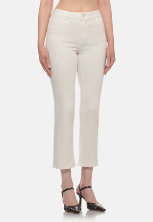 Donna che indossa pantaloni dritti color avorio tagliati sopra la caviglia e sandali neri con tacco su sfondo neutro.