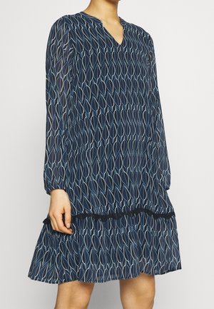 Femme portant une robe bleu marine à manches longues avec un motif feuille répétitif et des bordures noires au-dessus de l'ourlet à hauteur du genou.