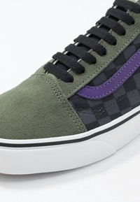 Gros plan d'une basket avec un bout en daim vert foncé, un tissu à carreaux noir et gris, une bande latérale violette, des lacets noirs et une semelle blanche avec une bande noire.