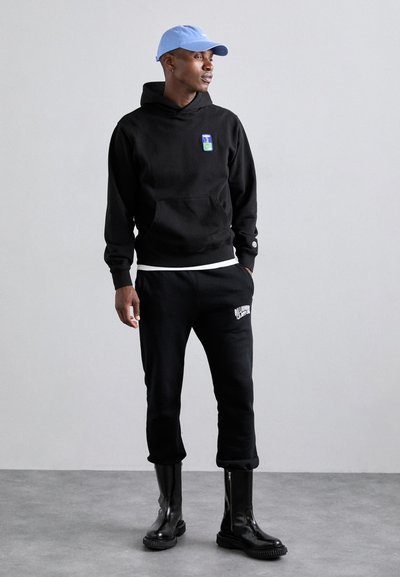 Felpa nera con cappuccio e tasca frontale, con logo, abbinata a pantaloni della tuta neri con testo bianco. Stivaletti neri lucidi alla caviglia. Cappellino azzurro chiaro.