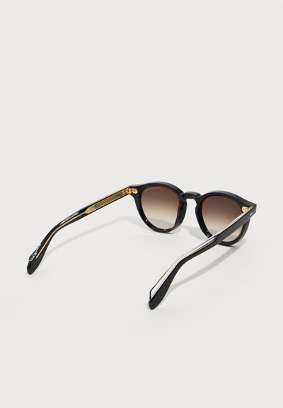 Μαύρα στρογγυλά γυαλιά ηλίου με καφέ φακούς διαβάθμισης και χρυσές λεπτομέρειες στα μπράτσα, τοποθετημένα σε λευκή επιφάνεια.