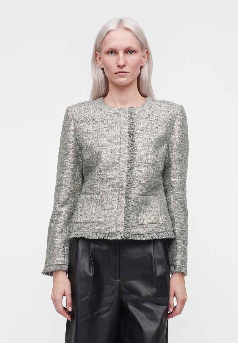 Jeune femme aux cheveux blonds raides porte une veste grise texturée à franges et un pantalon en cuir noir ample sur un fond uni.