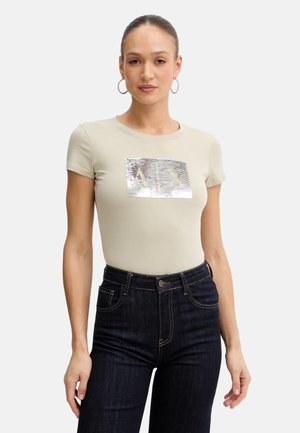 Femme avec des boucles d'oreilles créoles portant un t-shirt beige ajusté avec un motif rectangulaire argenté brillant et un jean taille haute bleu foncé.
