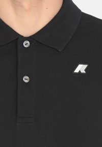 Polo shirt nera con colletto classico, placket con due bottoni e piccolo logo argento sul lato sinistro. Dettaglio del tessuto testurizzato visibile.