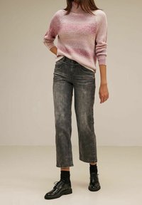 Personne portant un pull tricoté rose et blanc, un jean noir délavé coupe droite, des chaussettes noires et des chaussures à lacets noires sur un fond beige.