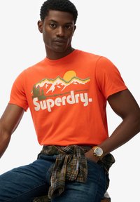 Orange bomulds T-shirt med bjerg- og solgrafik samt teksten "Superdry." Kortærmet, rund halsudskæring, parret med blå jeans og en ternet skjorte knyttet om taljen.
