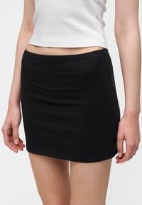 Femme portant une mini-jupe noire unie et un top court blanc, vue de la taille jusqu'au milieu des cuisses sur fond blanc.