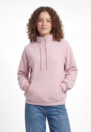 Giovane donna con capelli ricci che indossa una felpa rosa chiaro con zip a quarto e jeans blu, mani nella tasca frontale, guarda avanti.