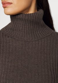 Brun ribbad polotröja med hög krage, som har ett strukturerat stickat mönster. Nära vy som fokuserar på material och struktur.
