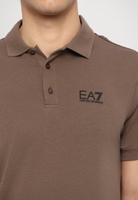 Hnědý polo tričko vyrobené z měkkého materiálu. Má límec, tři knoflíky a logo vytištěné na levé části hrudi v černé barvě.