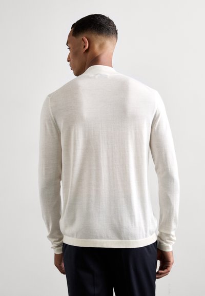 Uomo in piedi di spalle, vestito con un maglione bianco a maniche lunghe e pantaloni scuri, su uno sfondo chiaro e semplice.