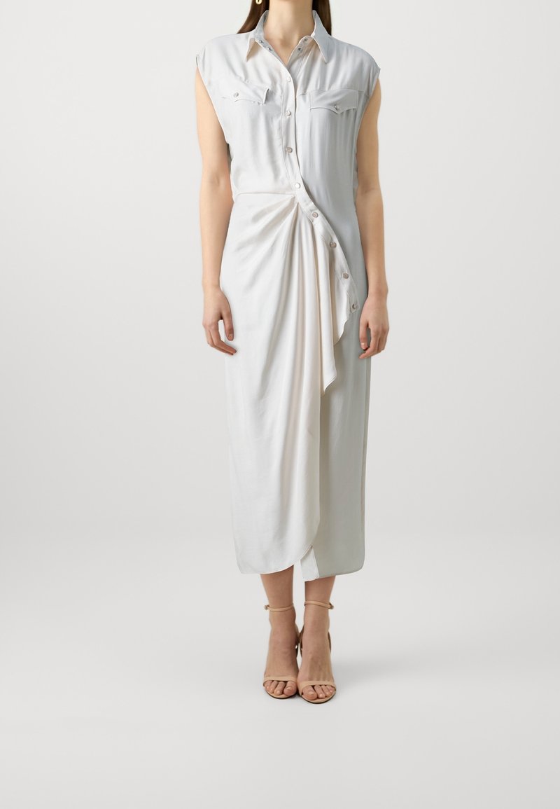 Robe blanche sans manches avec un col chemise, des boutons à l'avant et un détail de jupe drapée. Le tissu semble lisse avec un léger éclat ; longueur au genou.