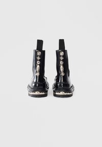 Botine din piele neagră, cu panouri elastice pe laterale. Accente metalice decorative pe partea exterioară și detalii texturate pe talpă.