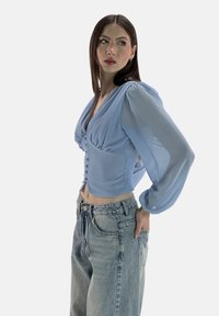 Helles blaues kurzes Blusenshirt mit Puffärmeln, einem V-Ausschnitt und Vorderknöpfen. Kombiniert mit hoch geschnittenen, vintage Denim-Jeans für einen Kontrast.