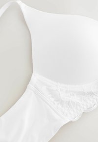 Reggiseno bianco con ferretto, caratterizzato da un tessuto morbido, rifiniture in pizzo lungo la banda e spalline regolabili. Forma curvilinea per supporto e copertura.