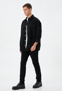 Camisa negra de gran tamaño con dos bolsillos frontales, botones plateados y puños remangados, combinada con pantalones negros ajustados y zapatos negros de suela gruesa.
