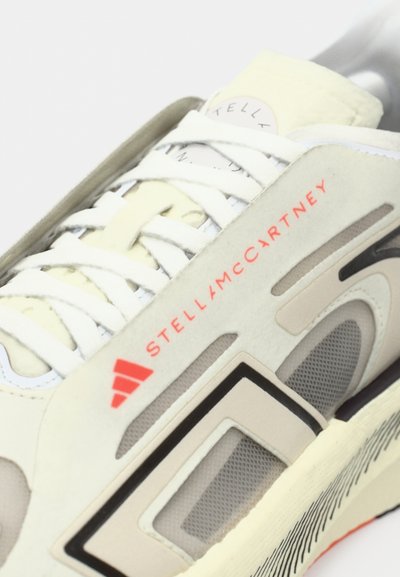 Białe i beżowe sneakersy z białymi sznurówkami, czerwonymi logo Stella McCartney i Adidas oraz siateczkową wstawką po boku.