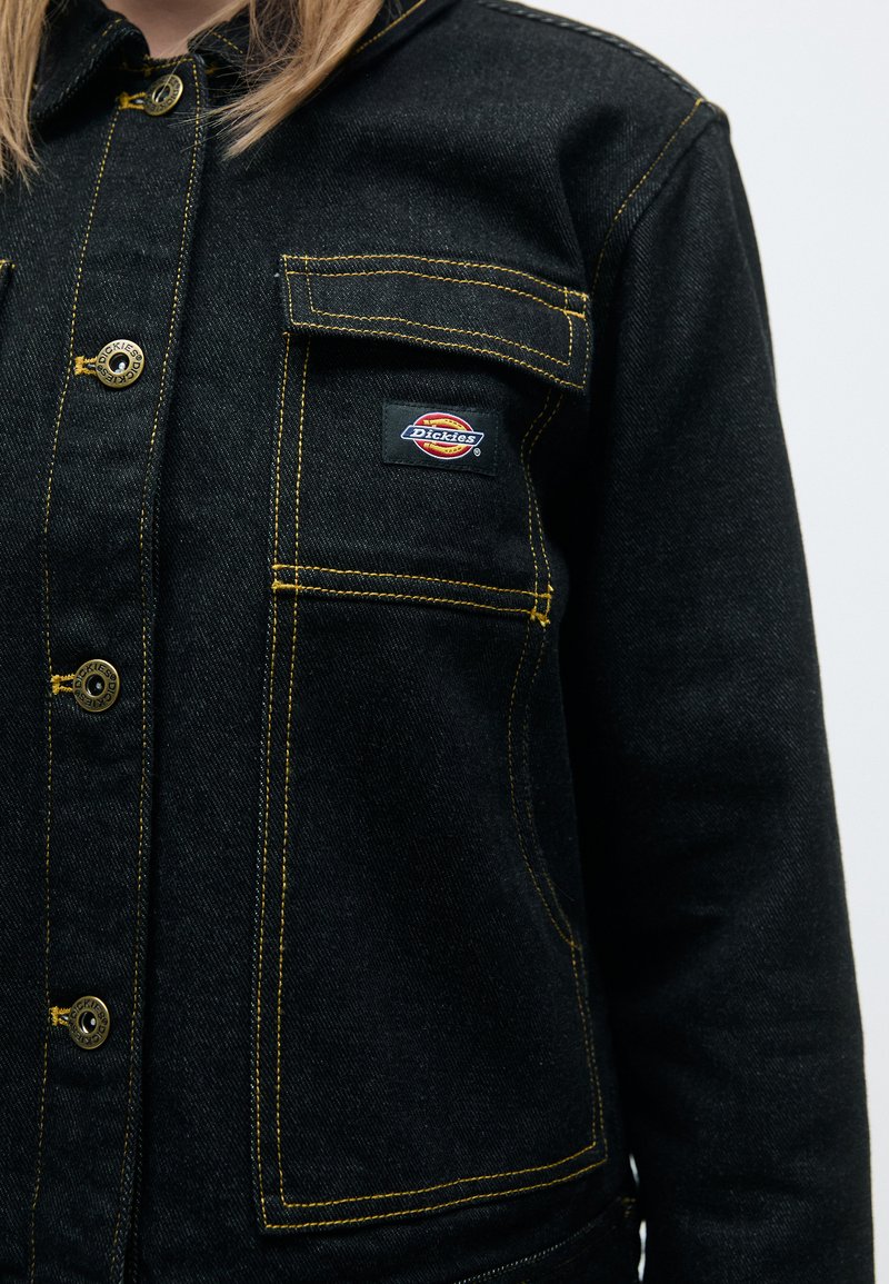 Chaqueta de mezclilla negra con costuras amarillas, cierres de botón y dos bolsillos en el pecho. Cuenta con un parche del logo de Dickies en el bolsillo izquierdo.