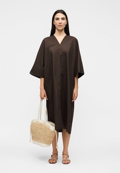 Γυναίκα με μακριά σκούρα μαλλιά που φοράει ένα φαρδύ σκούρο καφέ φόρεμα με V-λαιμόκοψη, κρατώντας ένα πλεκτό μπεζ τσάντα tote και φοράει καφέ σανδάλια.