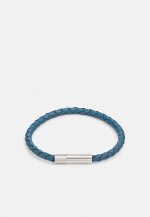 Bracelet tressé en cuir teal, avec un fermoir cylindrique en métal argenté. Texture lisse avec un motif de tressage complexe.