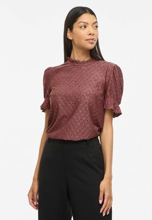 Kobieta ubrana w wrzosową, krótki rękaw, ażurową bluzkę z falbanianym kołnierzem oraz czarne spodnie z wysokim stanem, spogląda nieco na bok.