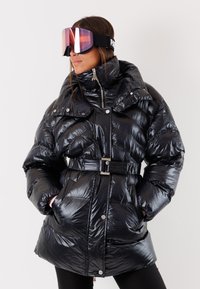 Giacca piumino nera con collo alto, vita con cintura, texture lucida, dettagli con bottoni e due tasche laterali. Modello con occhiali da sci.