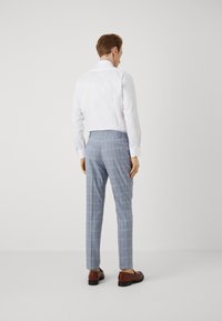 Cămașă albă cu guler și spate scurt, asortată cu pantaloni cu model carourile albastre deschise și pantofi brown slip-on, văzută din spate.