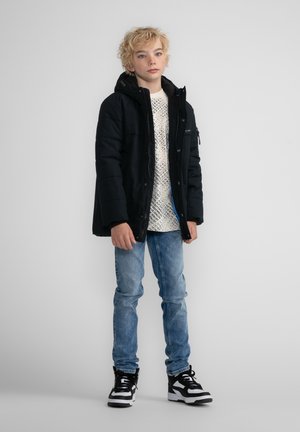 Giacca a vento nera con cappuccio, abbinata a un maglione bianco a fantasia e jeans azzurri chiari. Sneakers chunky in bianco e nero completano il look.